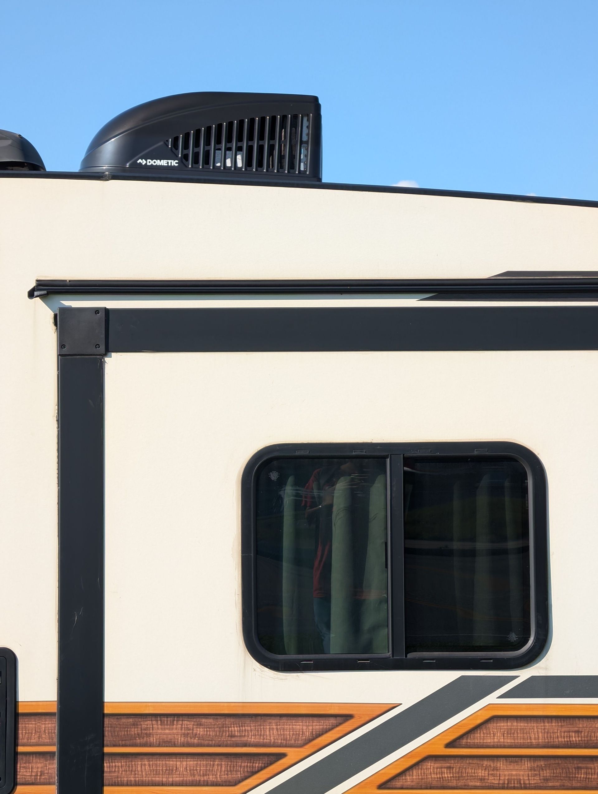 Side of a cream RV with black trim and a small tinted window under a blue sky
