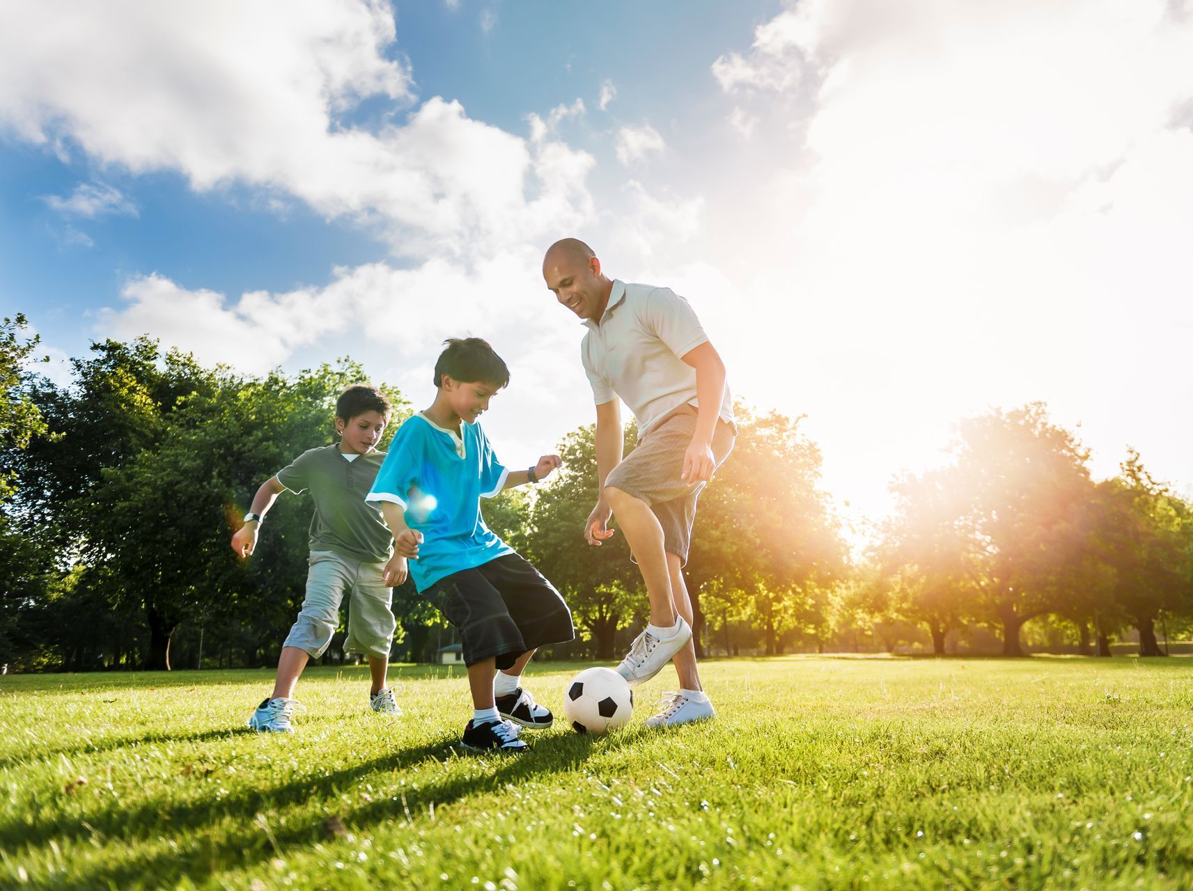 Father and two children playing soccer in a sunny park; all smiling and active on green grass.