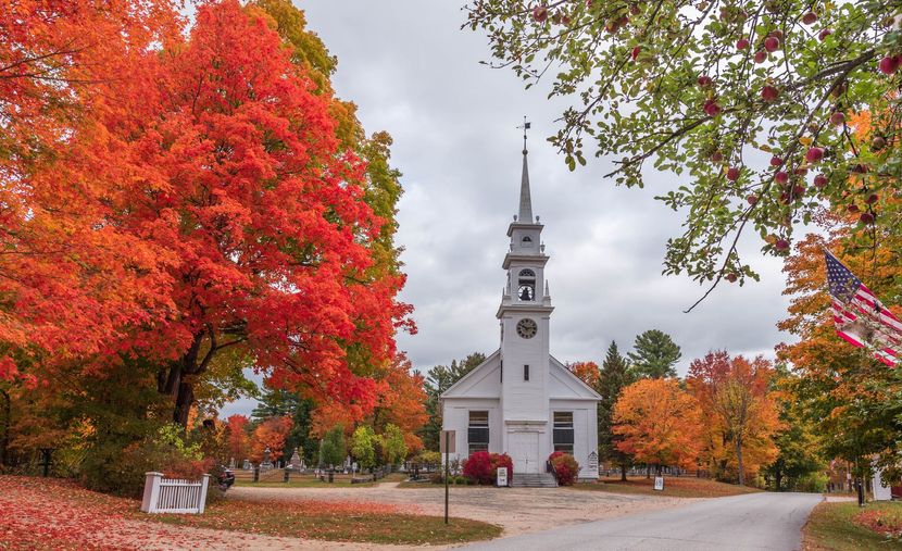 White church with steeple on a tree-lined road, framed by vivid red autumn foliage.