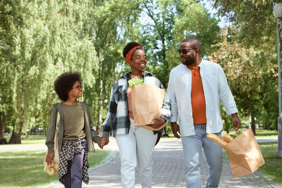 Three people walking in a sunny park, smiling and carrying shopping bags.