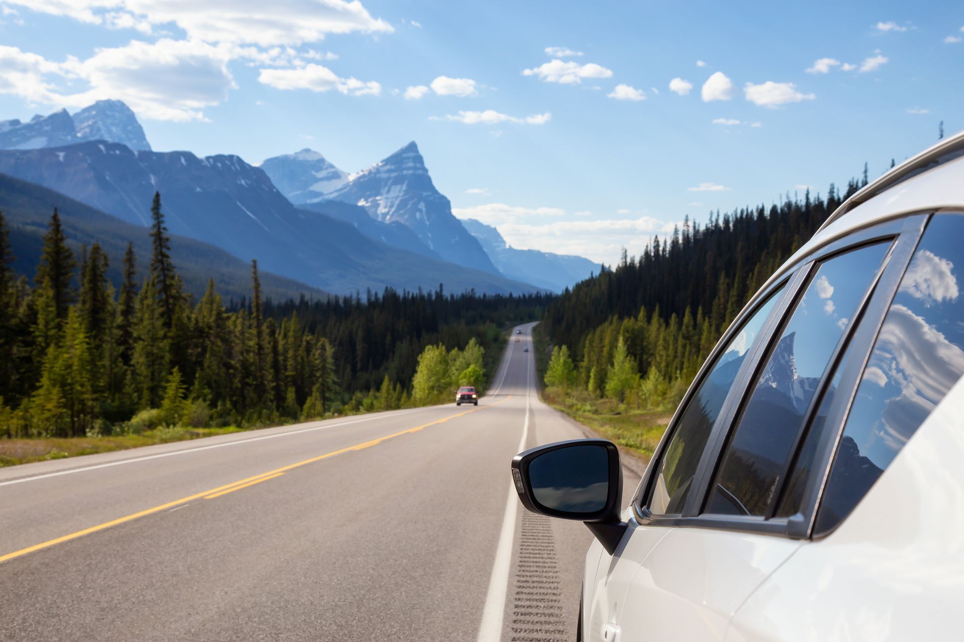 White car on an open mountain road with pine trees and snow-capped peaks under a blue sky