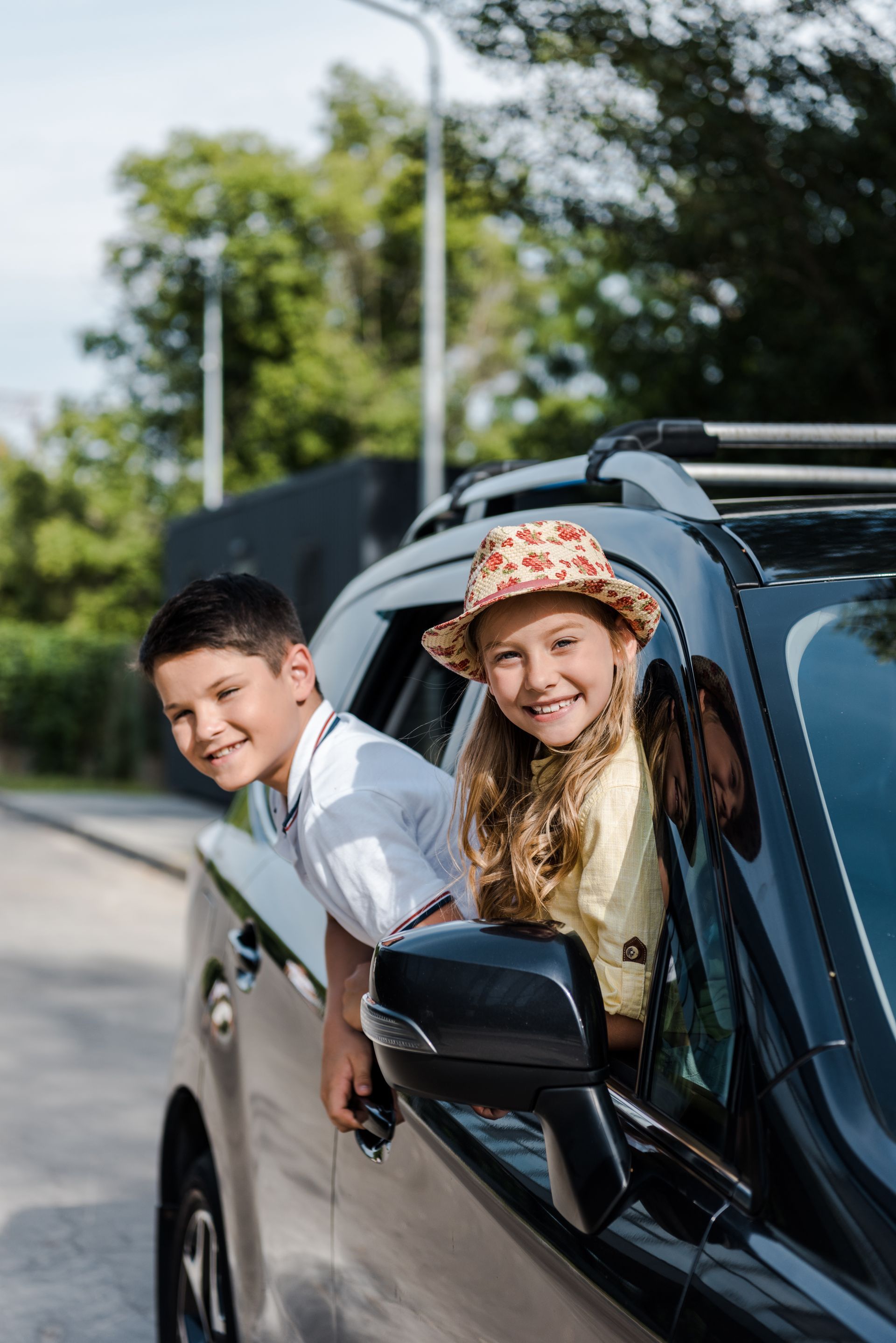 Two children lean out of a car window, smiling on a sunny road