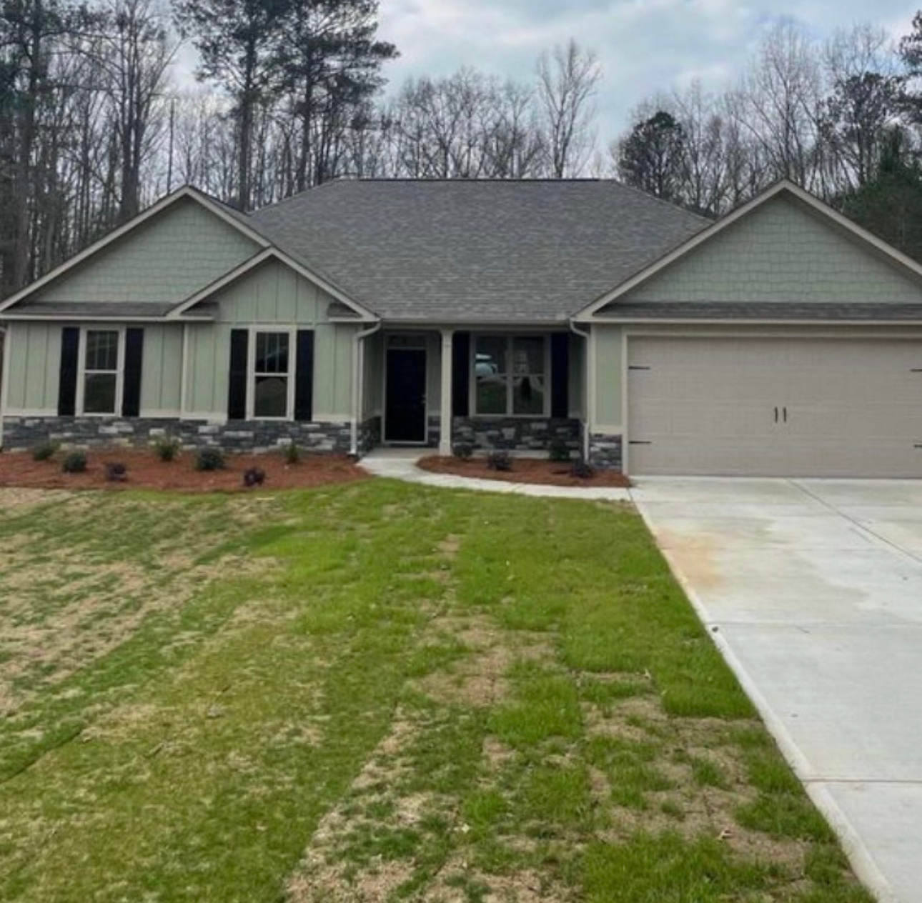 Green and gray ranch-style house with a driveway, small yard, and forest background.
