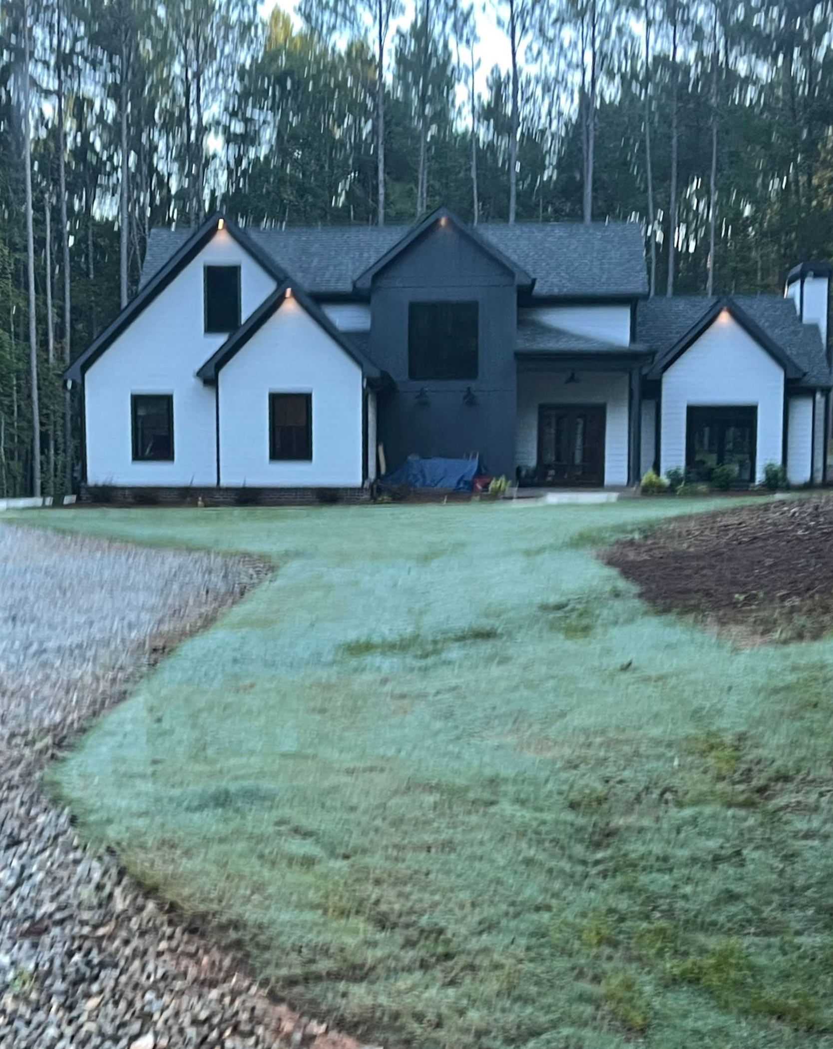 White house with dark trim, gravel driveway, and green lawn surrounded by trees.