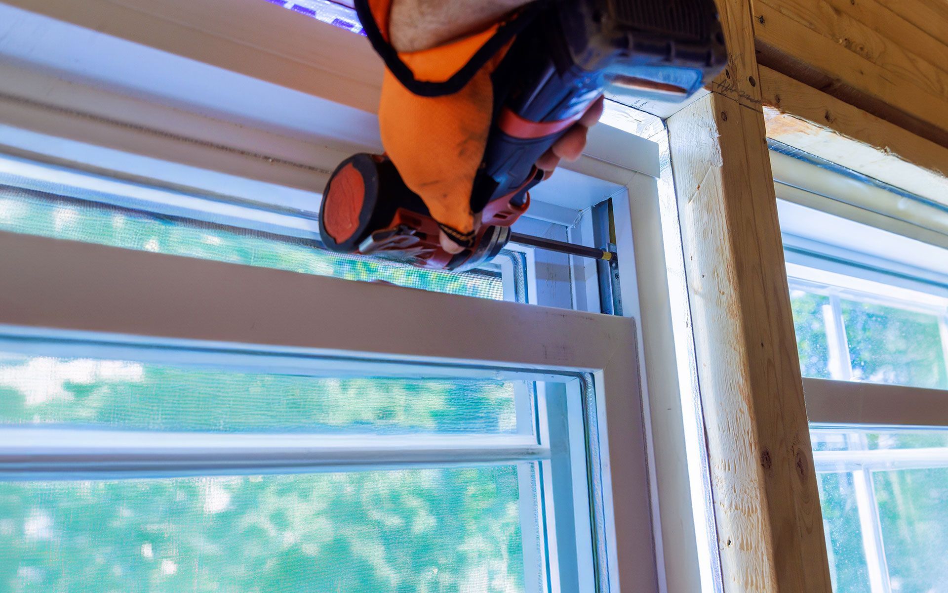 Person in orange work gloves using a drill to install a white window in a wooden frame.