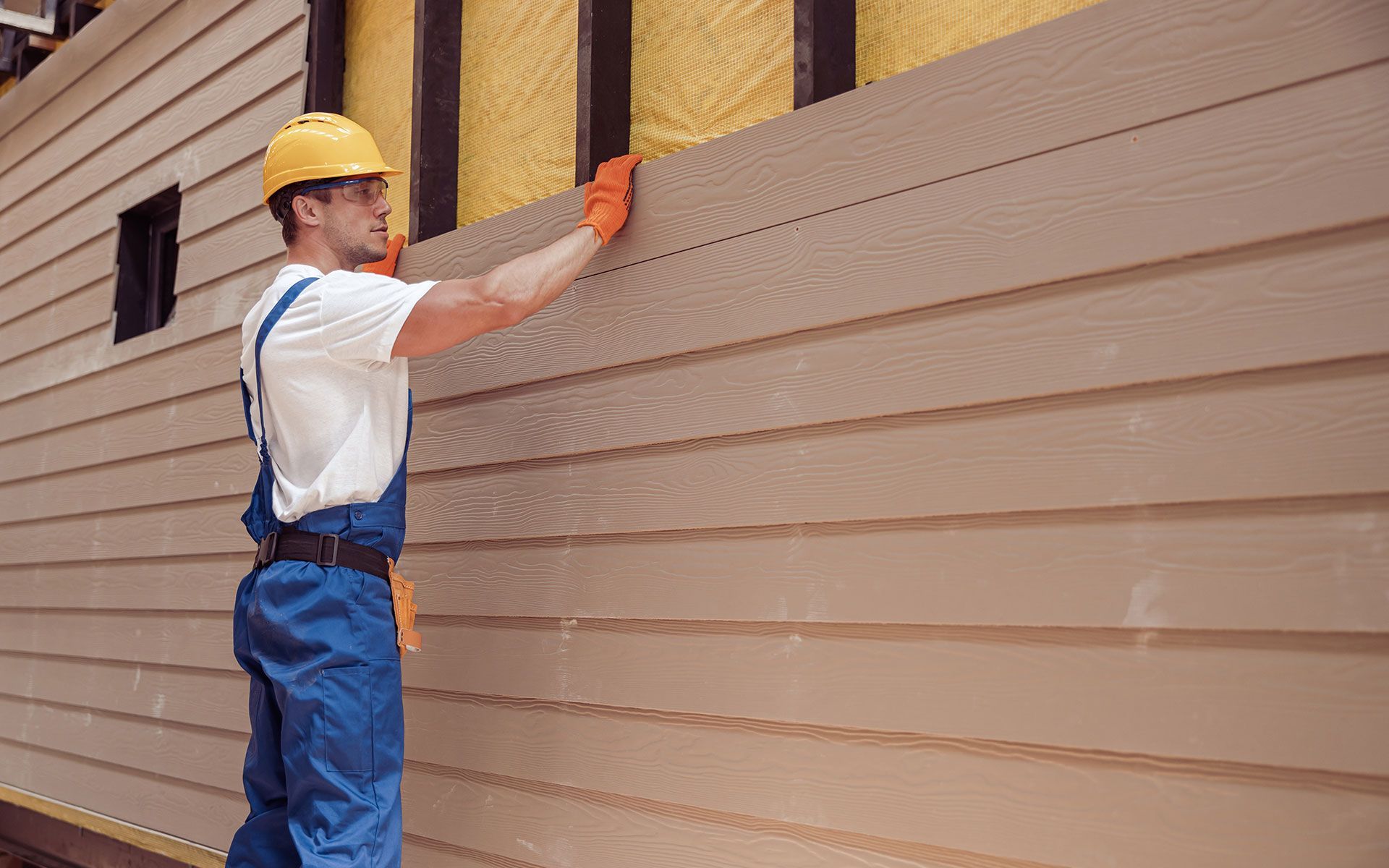 Construction worker in blue overalls and hard hat installing siding on a building.