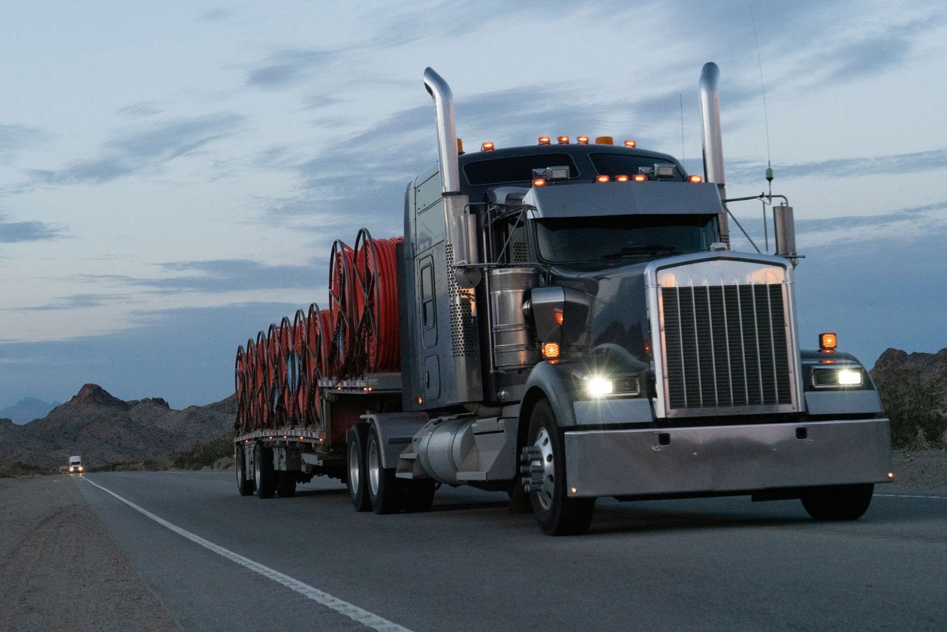 Semi-truck hauling large cable spools on flatbed trailer at dusk on highway.