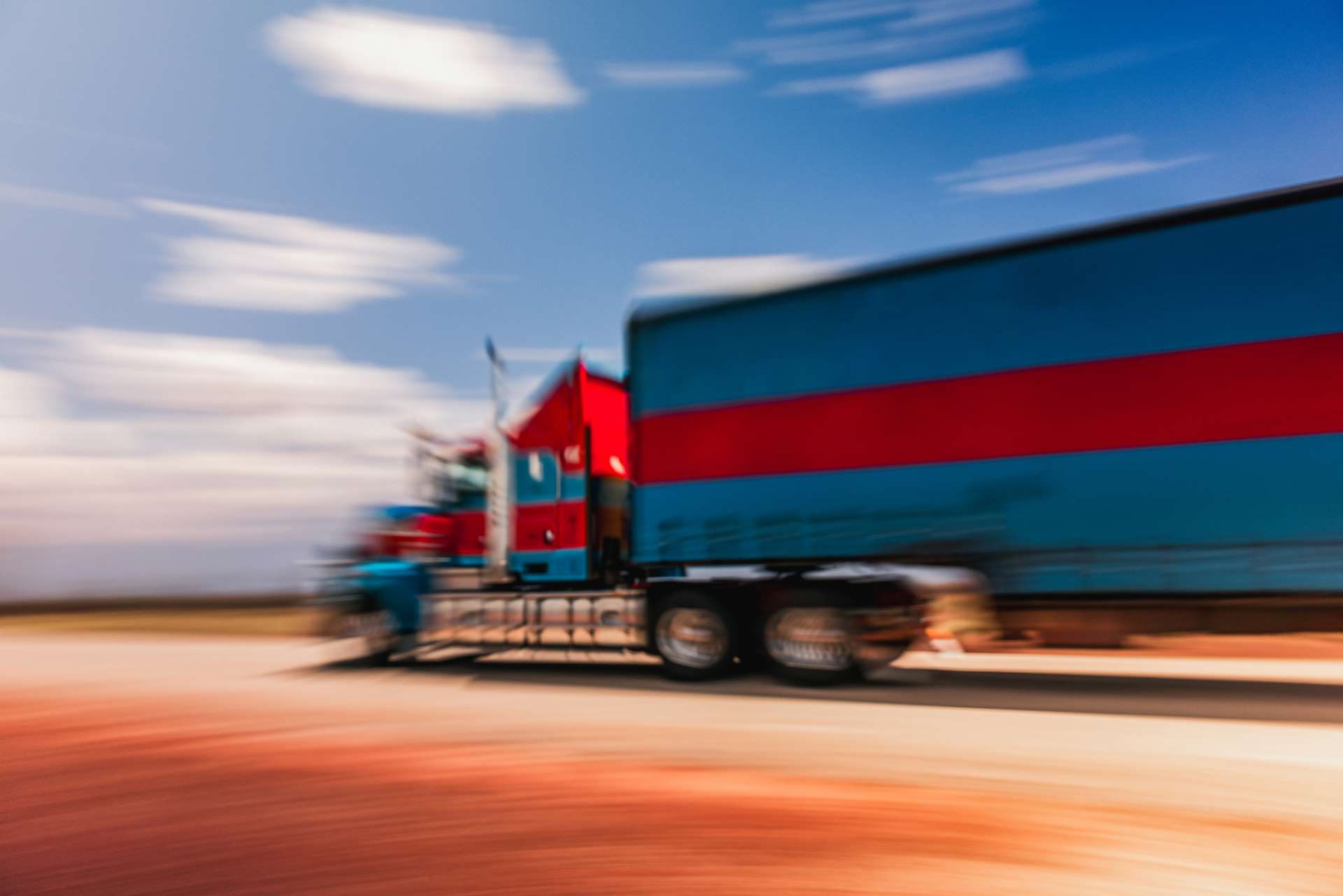 Blurred semi-truck with blue and red trailer driving under a partly cloudy sky.