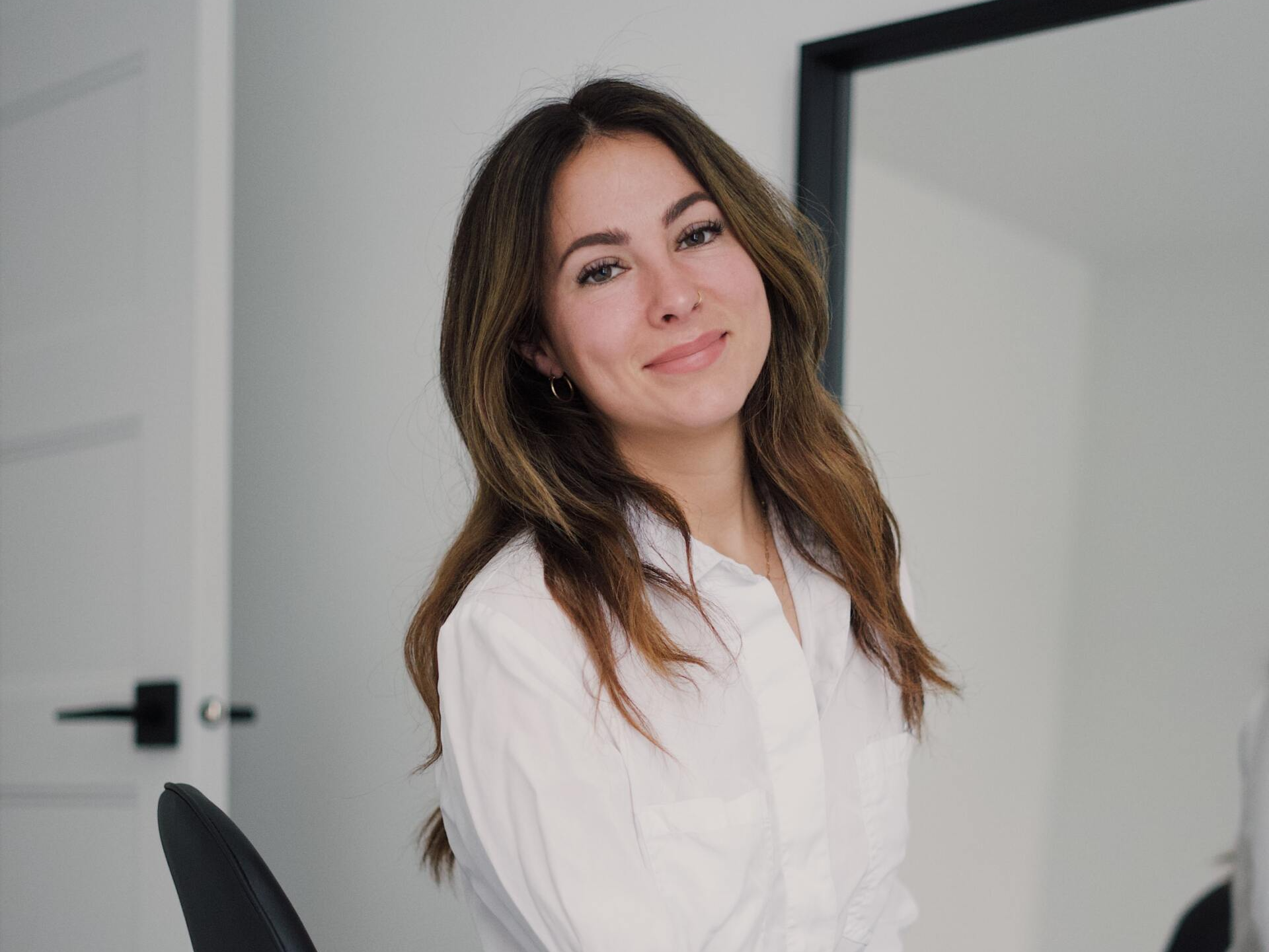 Una mujer con una camisa blanca está sentada en una silla y sonriendo a la cámara.