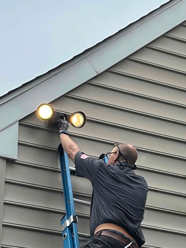 A man is standing on a ladder fixing a light on the side of a house.