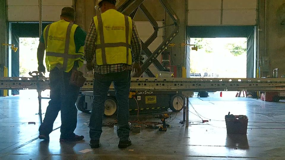 Two men in yellow vests are working on a machine in a warehouse.