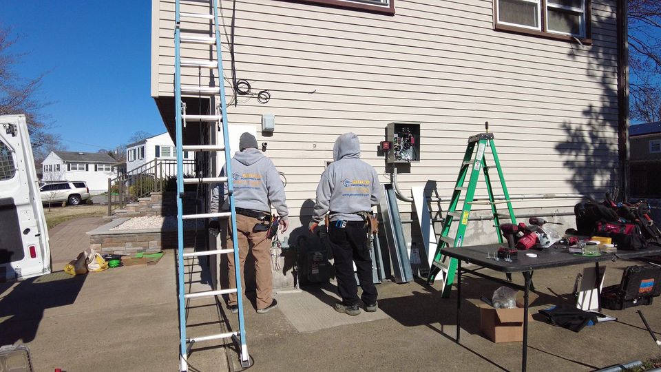 Workers on a ladder and scaffold repairing a house exterior, with tools and supplies on the driveway.
