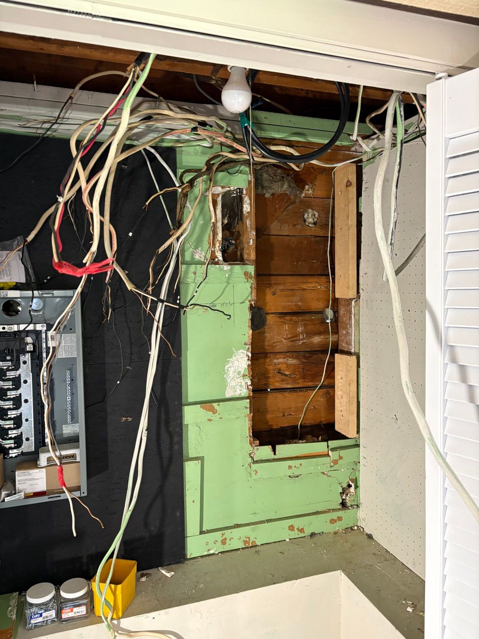 Open electrical panel with exposed wiring and damaged green wall inside a utility space