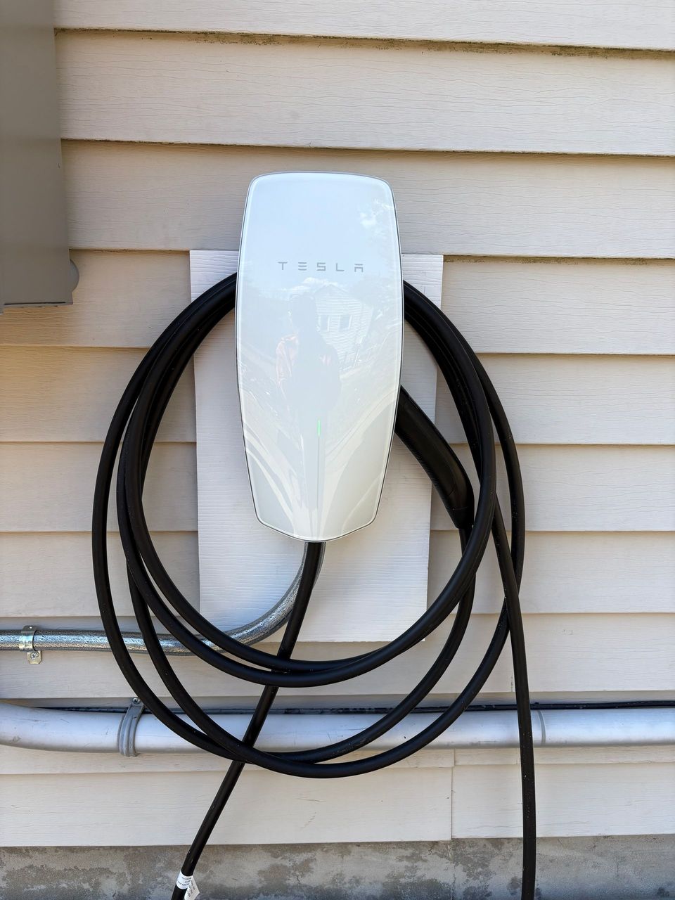 Wall-mounted white EV charging unit with black cable coiled against beige house siding