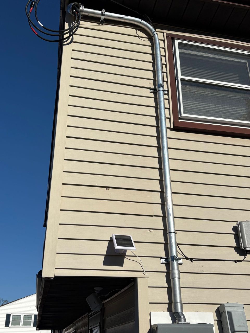 Exterior side of a beige house with white trim, downspout, and utility lines against a blue sky