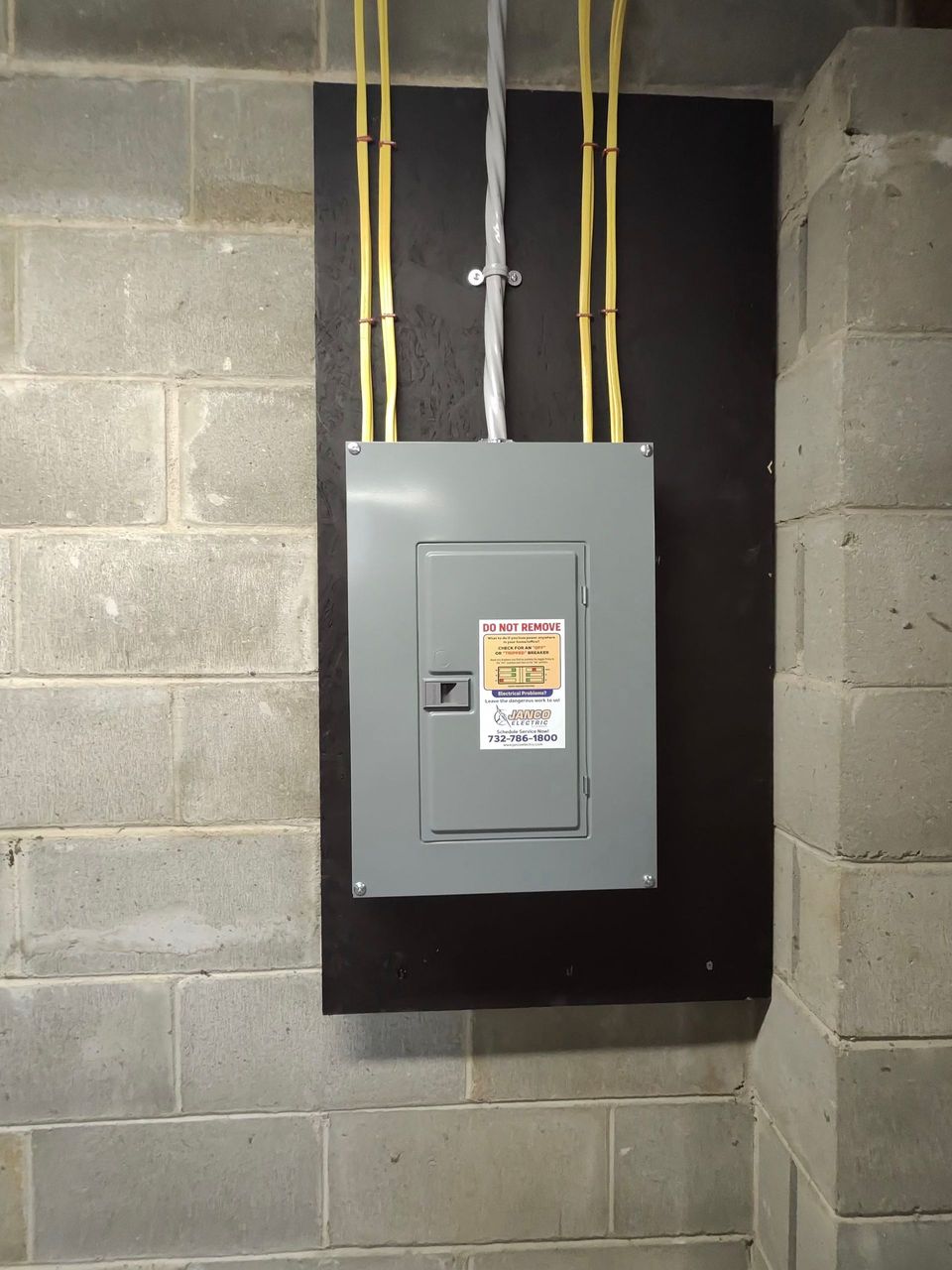 Gray electrical breaker panel mounted on a cinder block wall with yellow and white wires above it