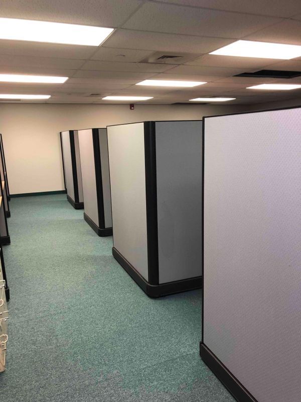 Empty office cubicles with gray partitions and teal carpet under fluorescent ceiling lights