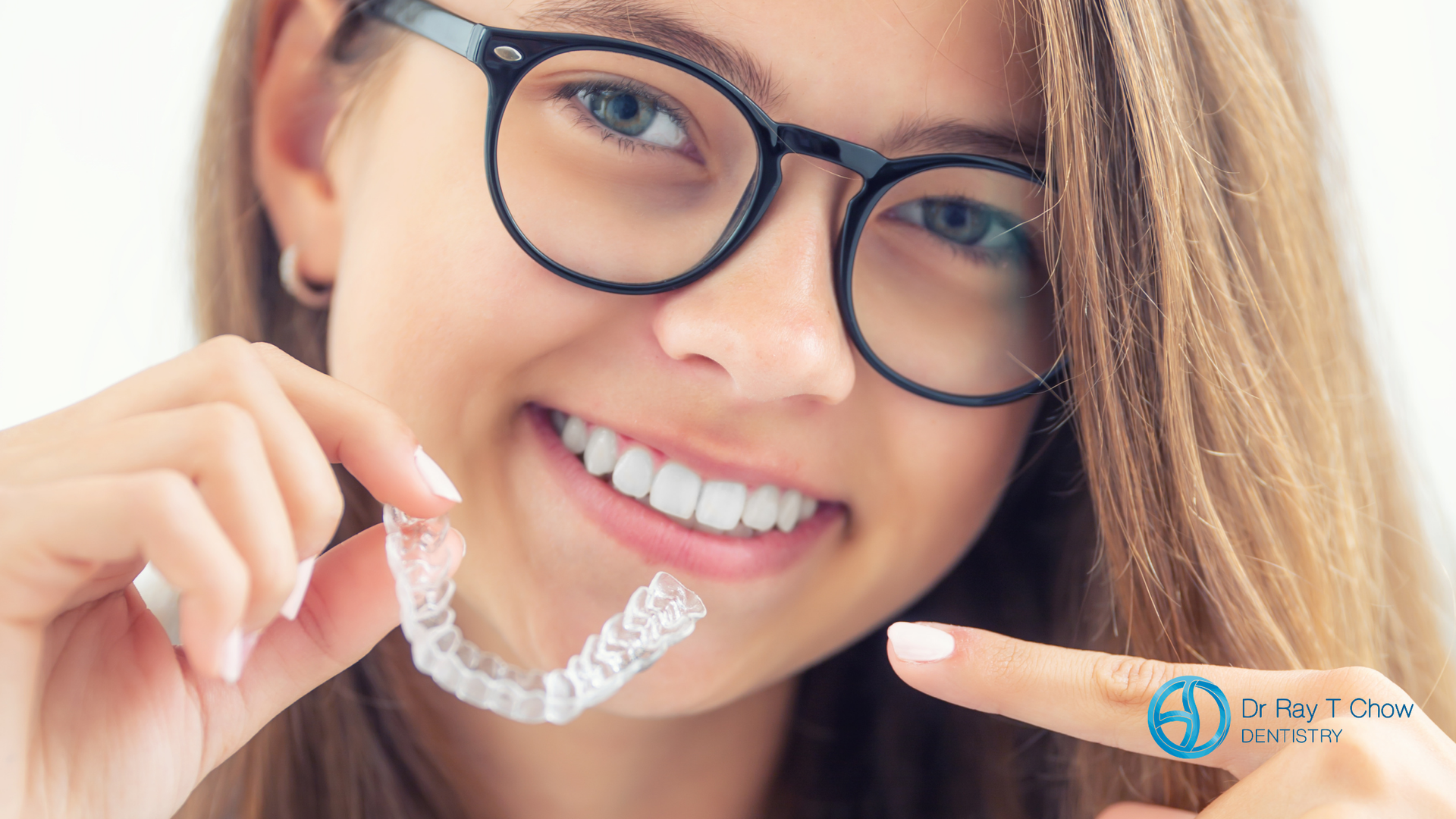 A young woman wearing glasses is holding a clear retainer in her mouth.
