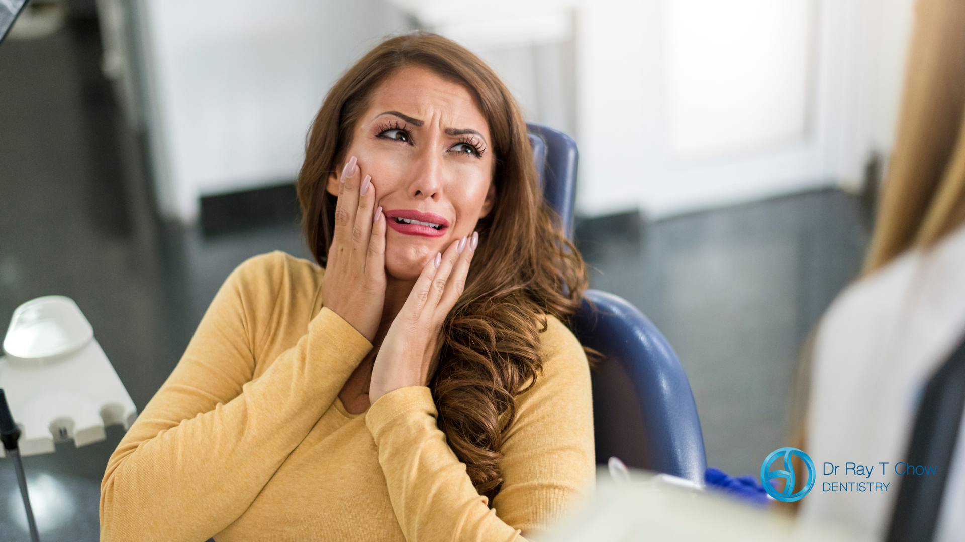A woman is sitting in a dental chair with her hands on her face.