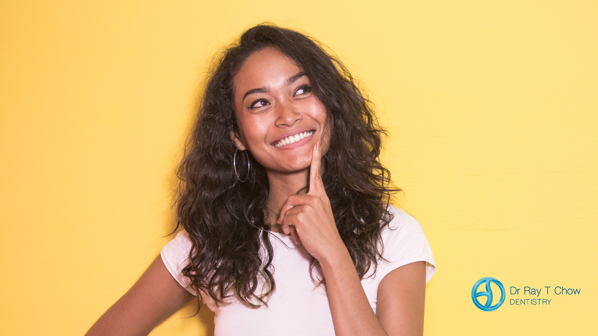 A woman is smiling and looking up while holding her hand to her chin.