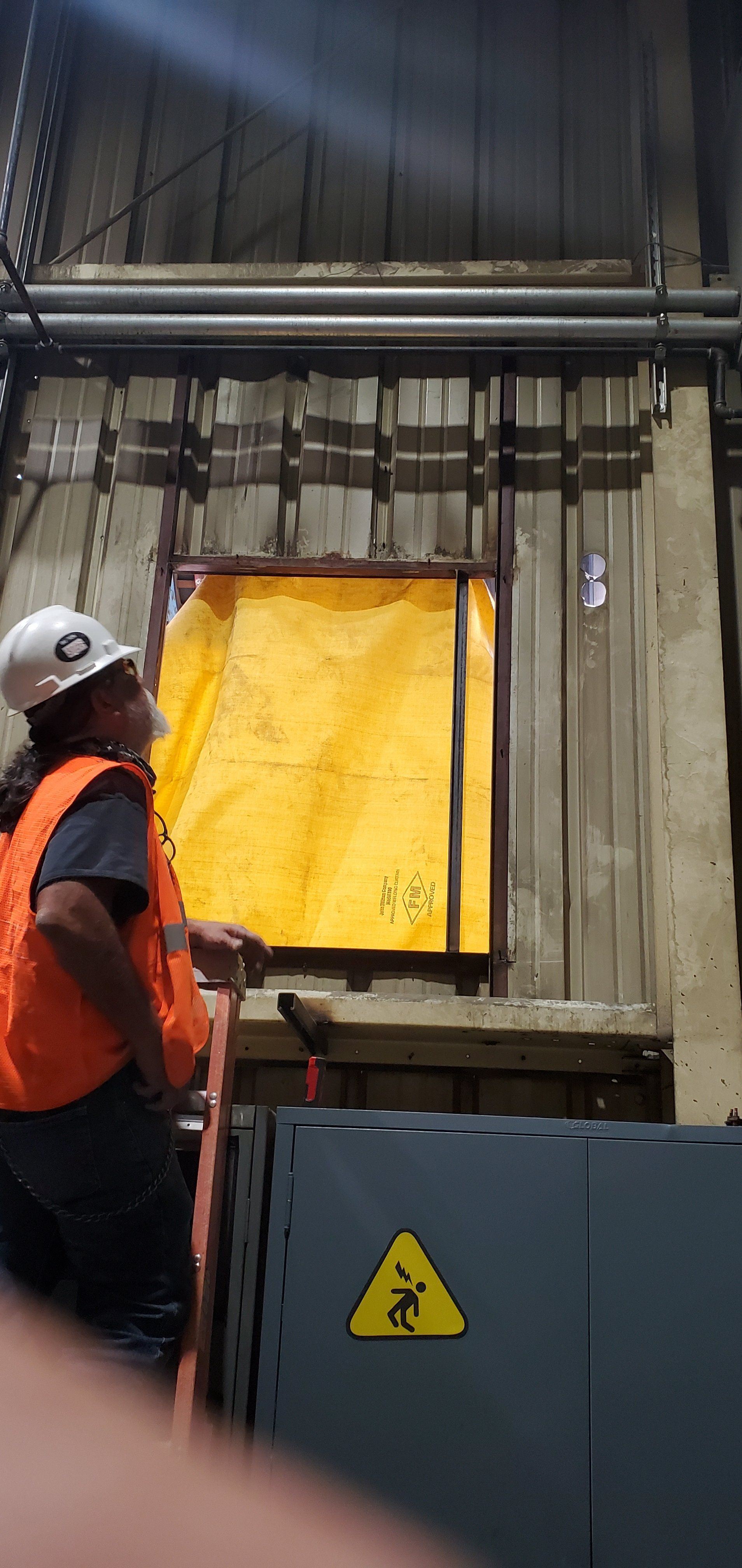 Worker in hard hat and safety vest, looking up at a yellow sheet, next to a hazard warning sign.