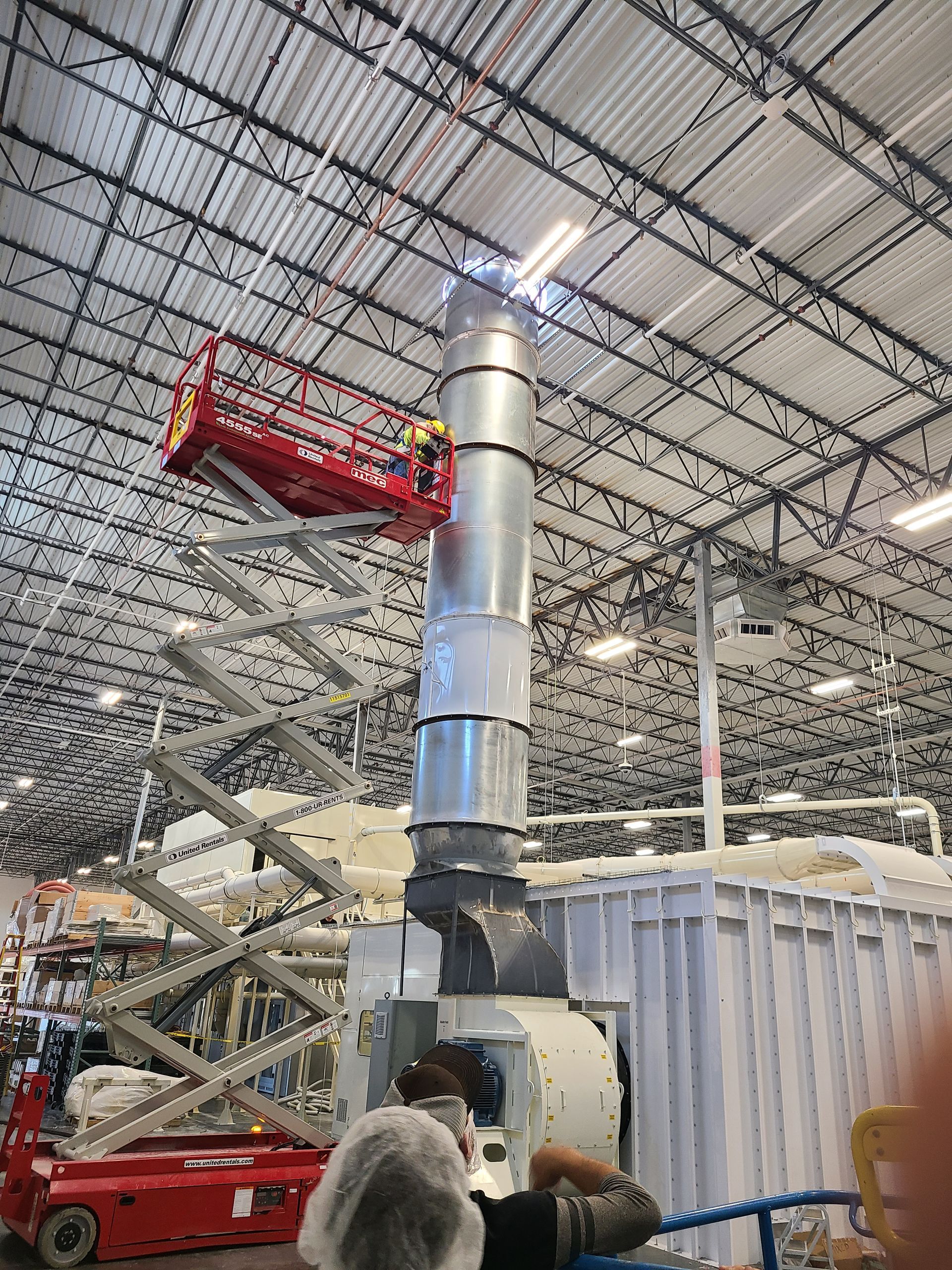 Workers on a lift welding ductwork inside a warehouse. Silver metal, red lift, blue railing.