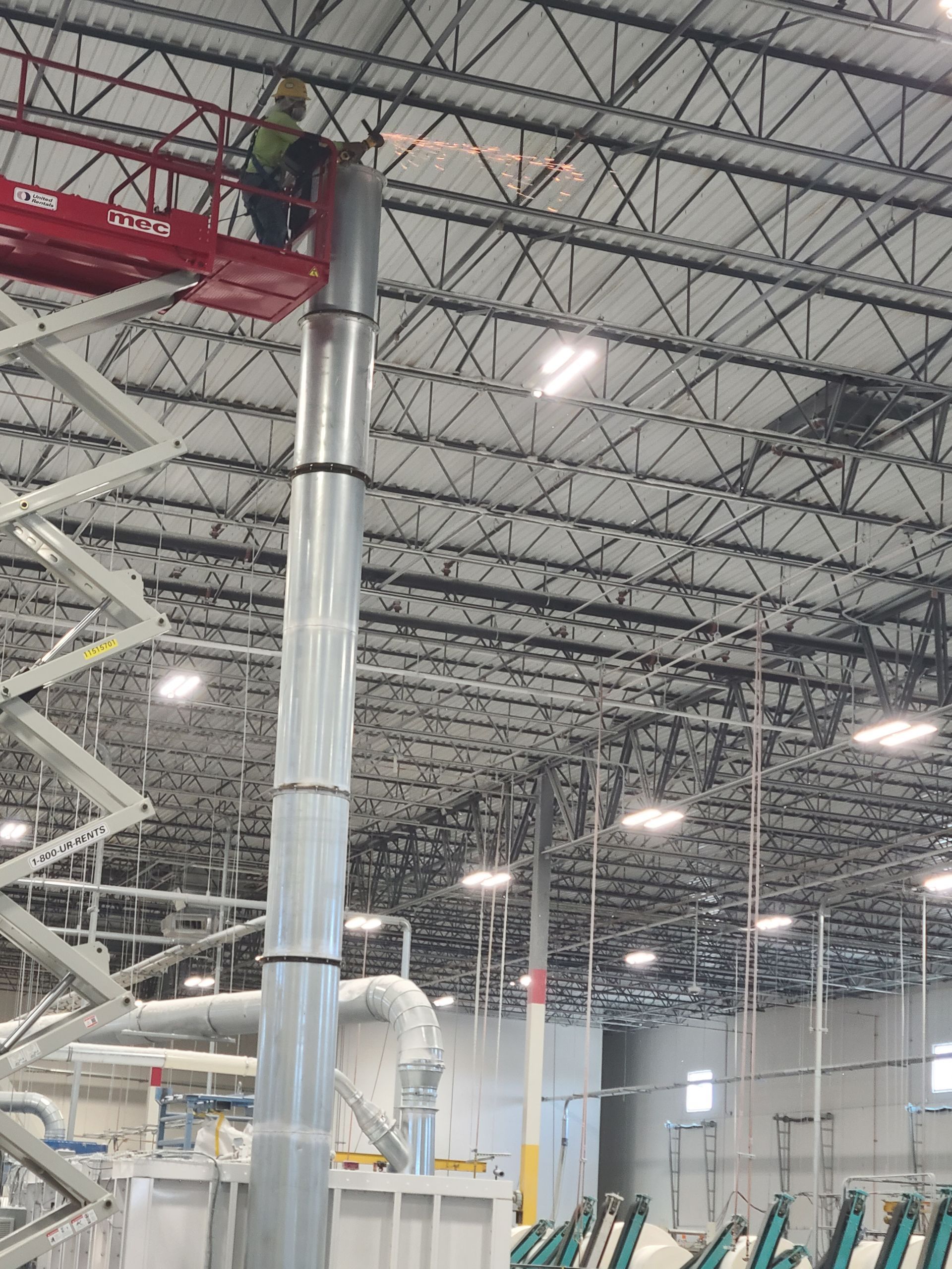 A worker in a lift welds ductwork inside a large industrial building.