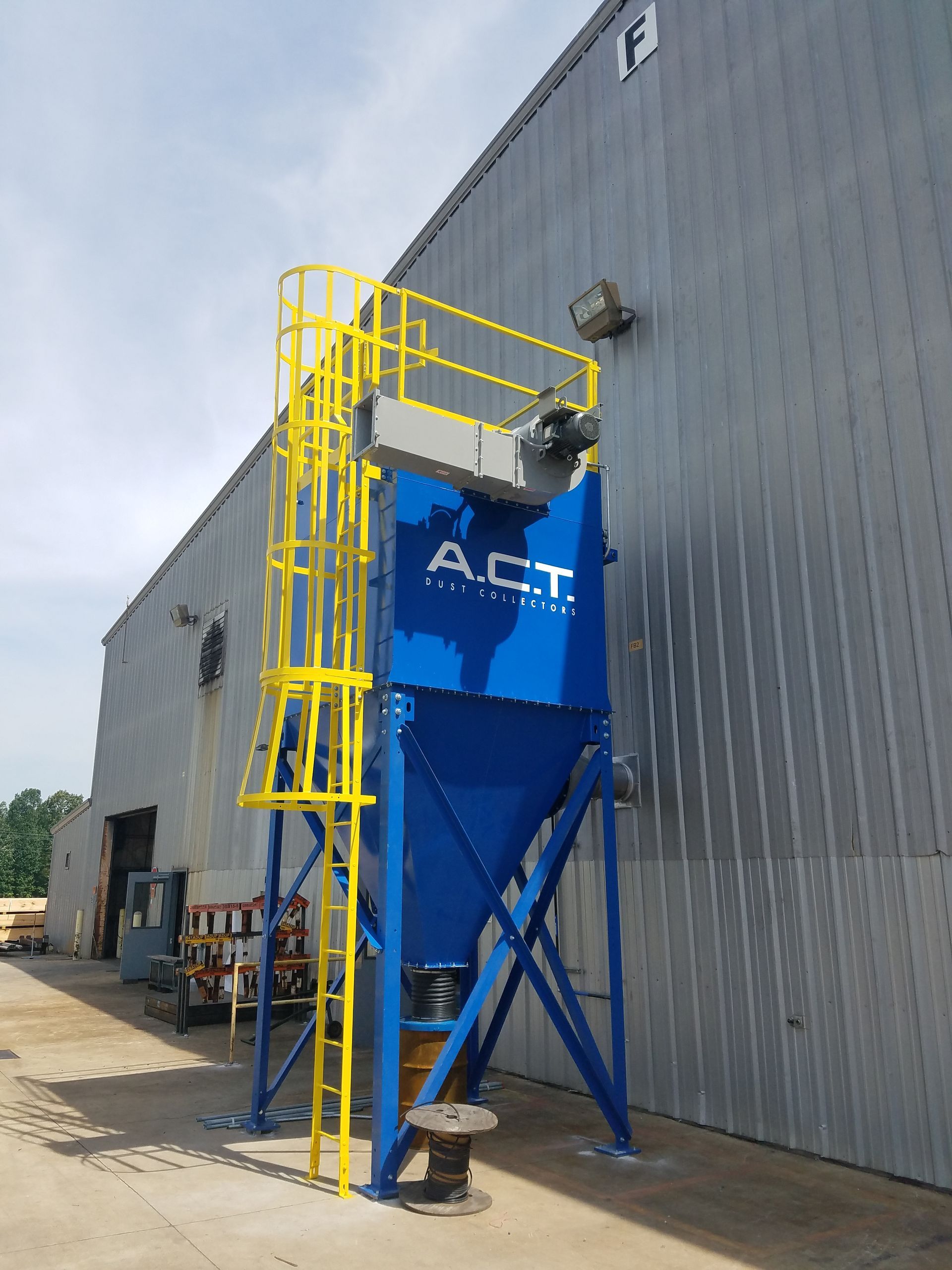 Blue industrial dust collector with yellow ladder against a gray building, outside.