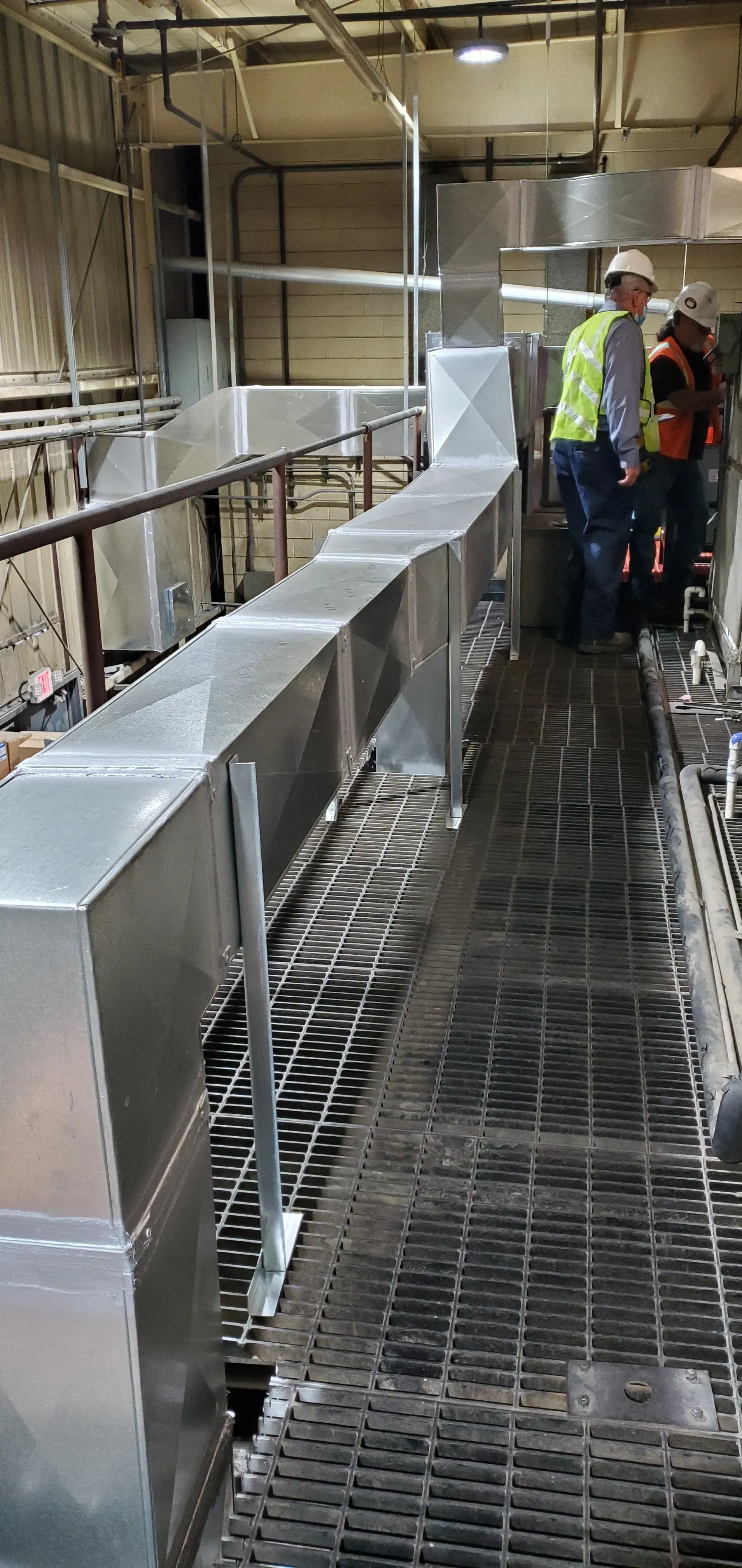 Workers standing near an industrial ventilation duct on a metal grate walkway.