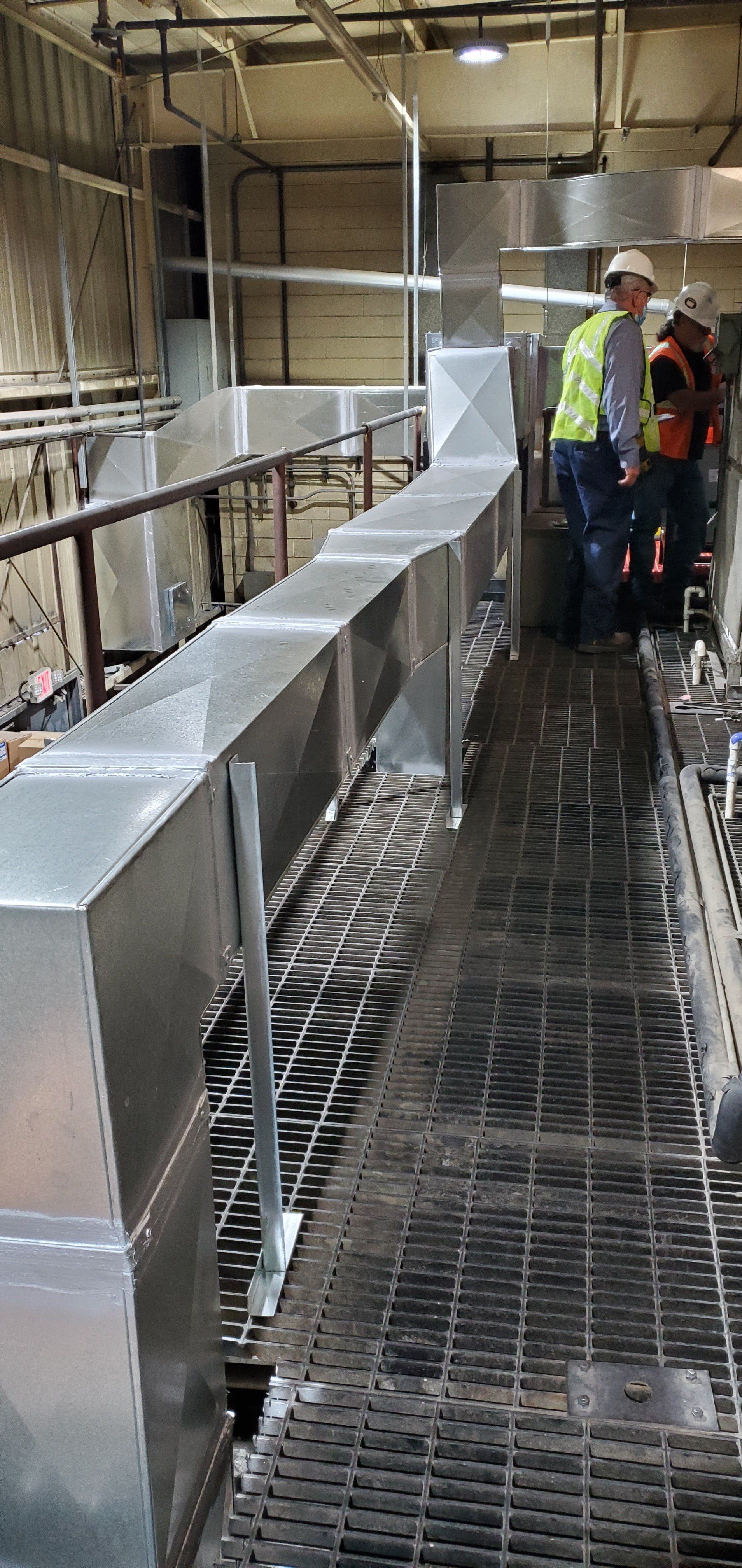 Two workers near rectangular metal ductwork on a metal grate walkway in an industrial setting.
