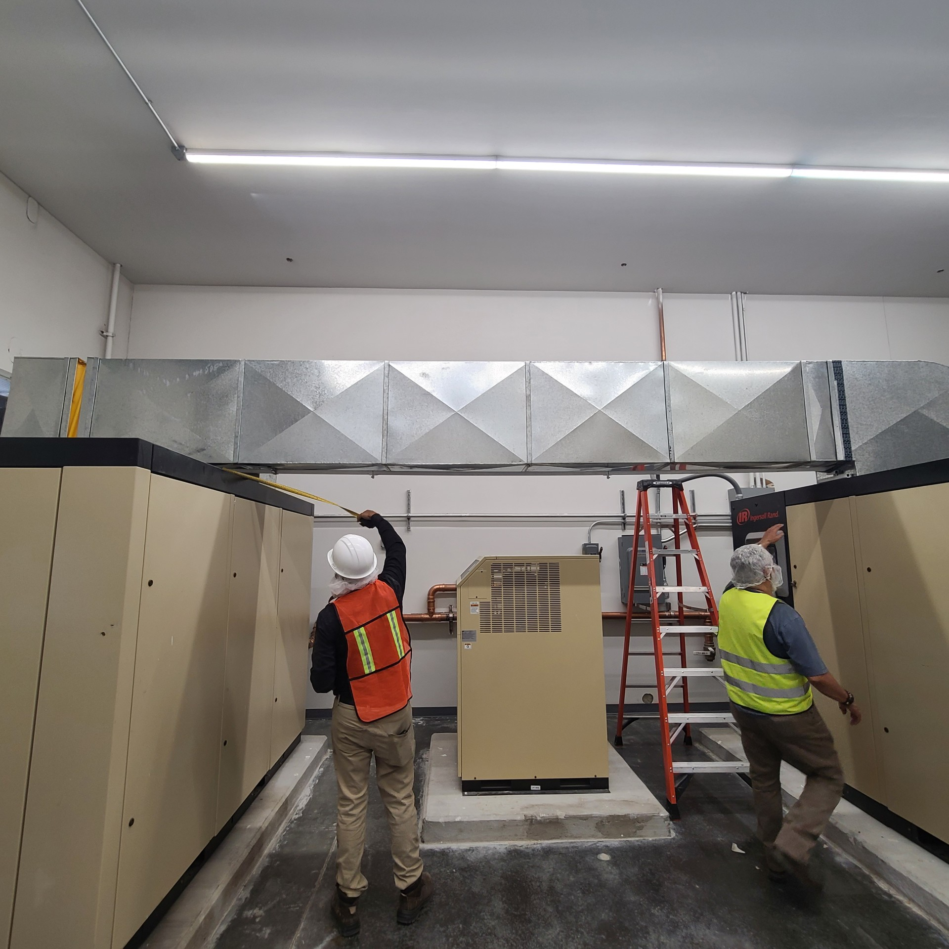 Two workers install ductwork in a beige equipment room; one measures, the other assists.