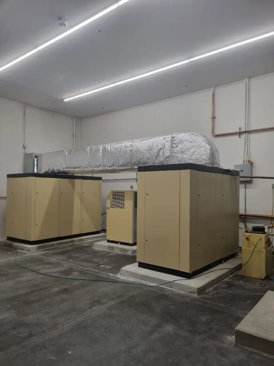 Three beige industrial machines in a white room with ductwork and piping.
