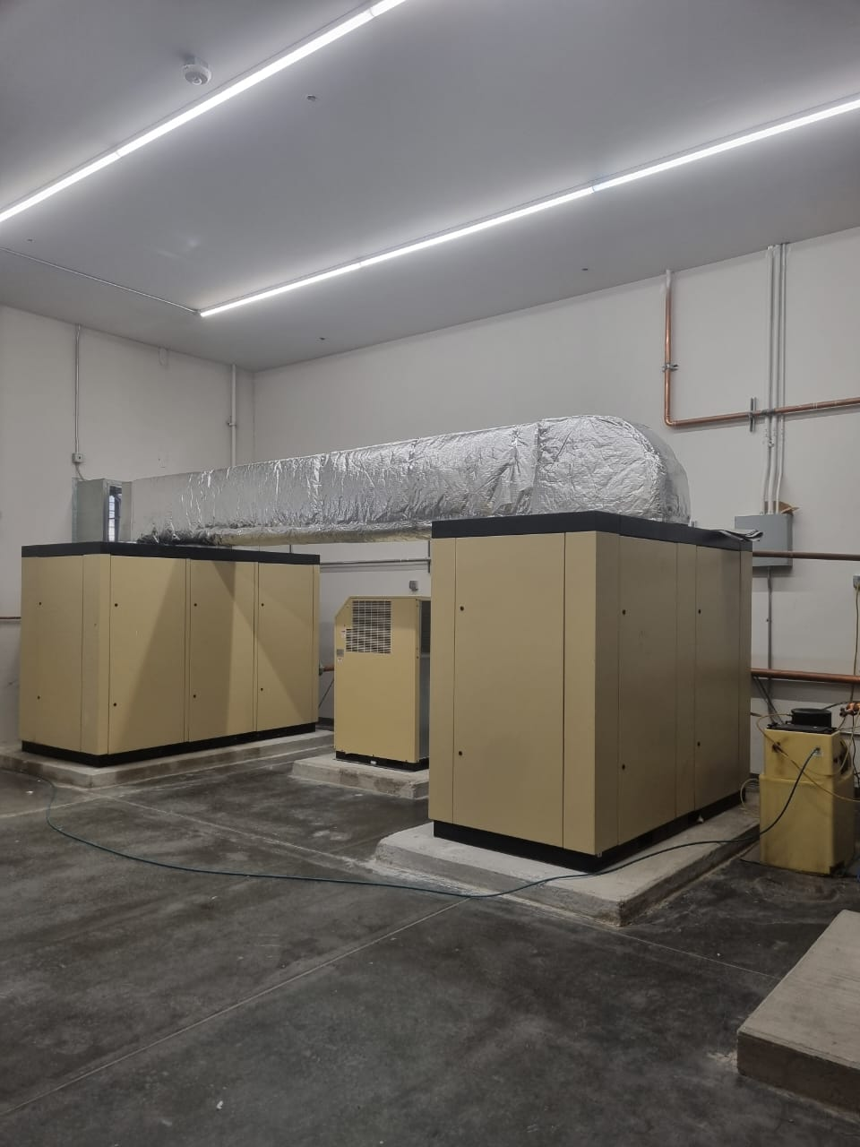 Three beige industrial machines on concrete bases in a white room; ductwork overhead.