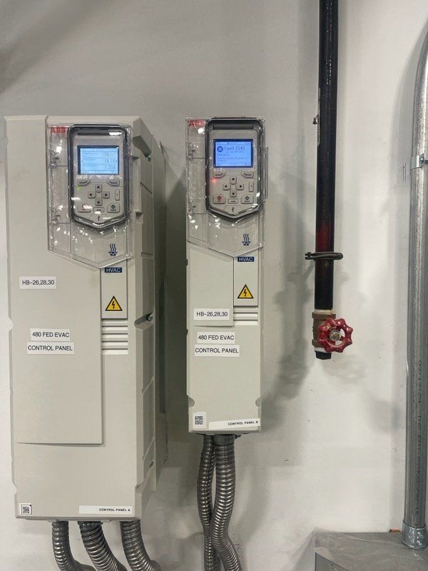 Two light gray industrial control units mounted on a white wall, display screens lit up, near a black pipe and red valve.