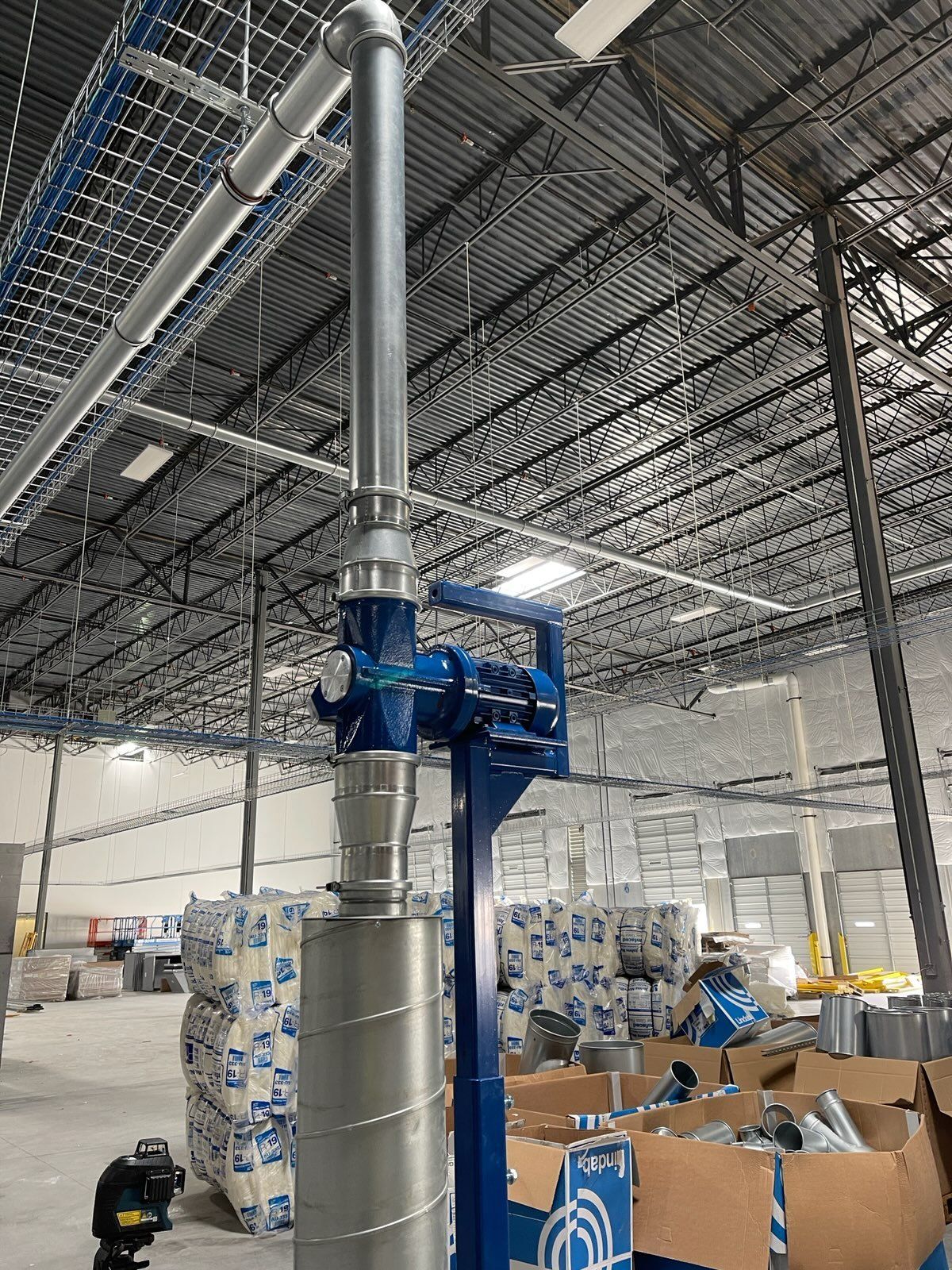 Blue and silver industrial ventilation system in a warehouse, with stacks of items in the background.
