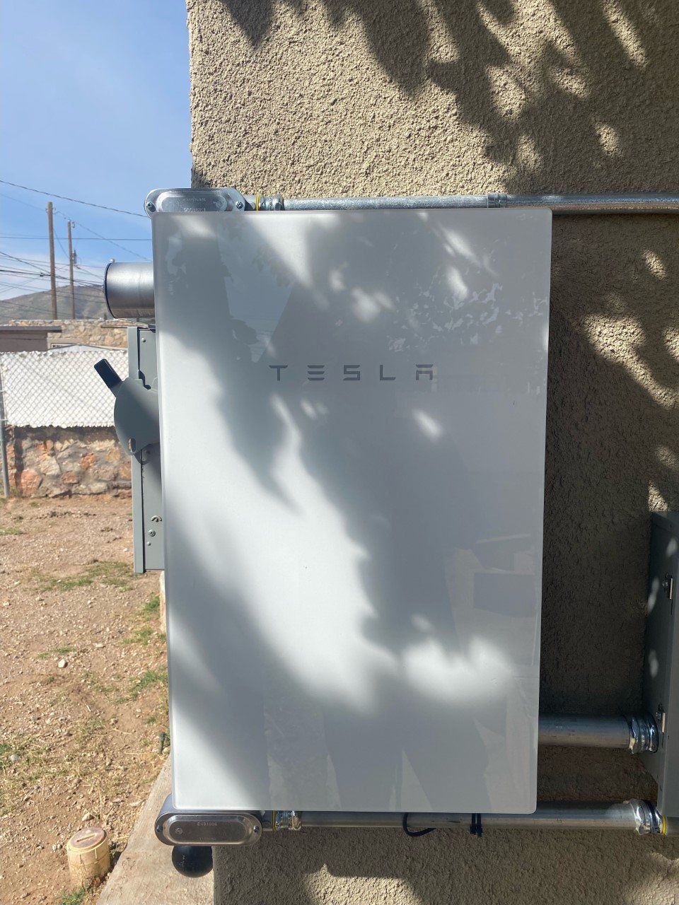 White Tesla Powerwall mounted on an exterior wall with conduit.