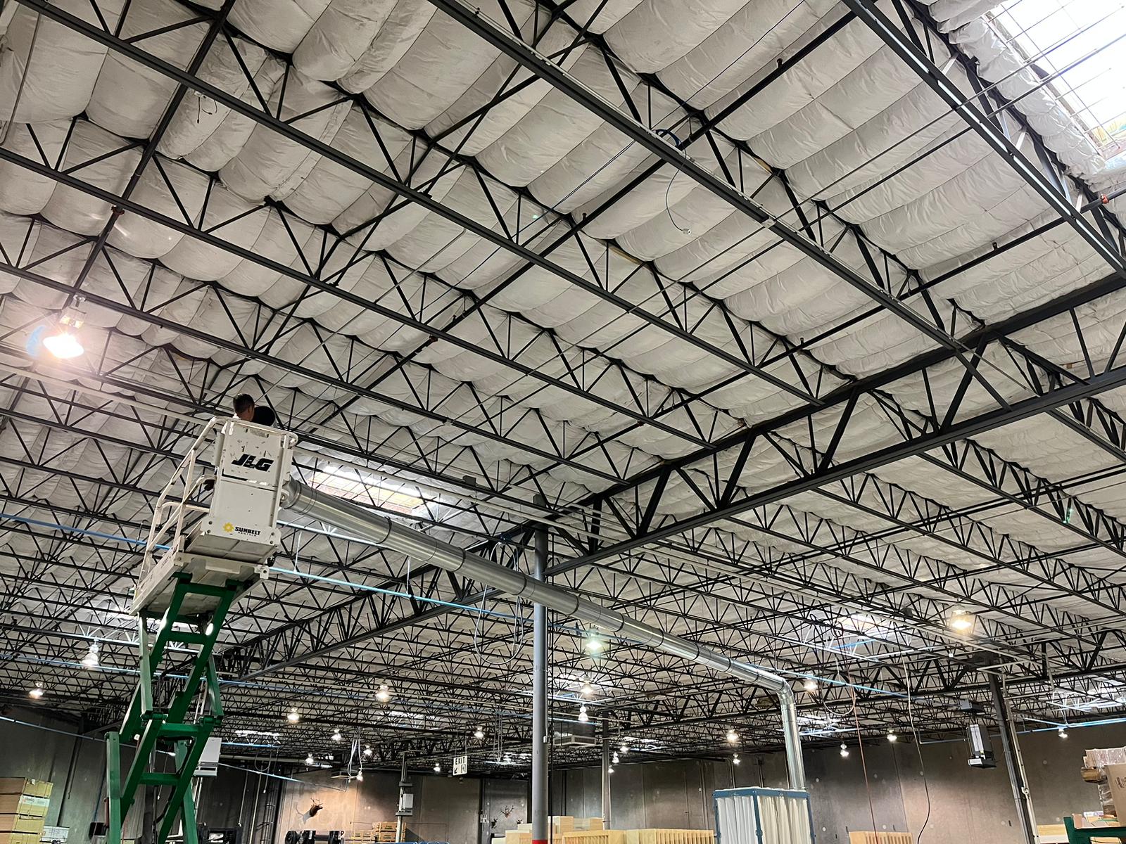 Warehouse ceiling with metal trusses and a person on a lift; overhead lighting.