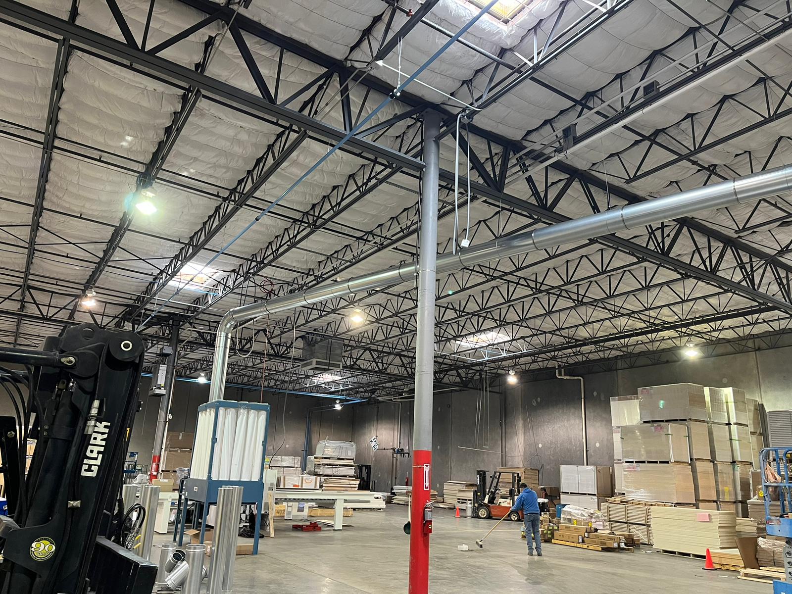 Warehouse interior with overhead beams, skylights, and a person walking. Forklift and stacked boxes are present.