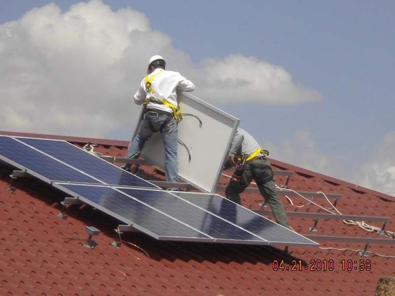 Two workers installing a solar panel on a red tile roof, wearing safety harnesses.