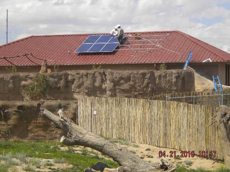 Person installing solar panels on a red-roofed building. A wooden fence and tree are in the foreground.