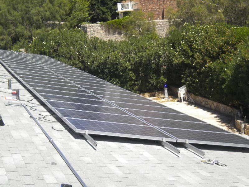 Solar panels installed on a rooftop. Dark panels, gray roof, greenery in the background.