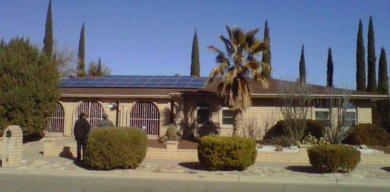 House with solar panels on the roof, palm tree, and hedges in front. Person standing near a hedge.