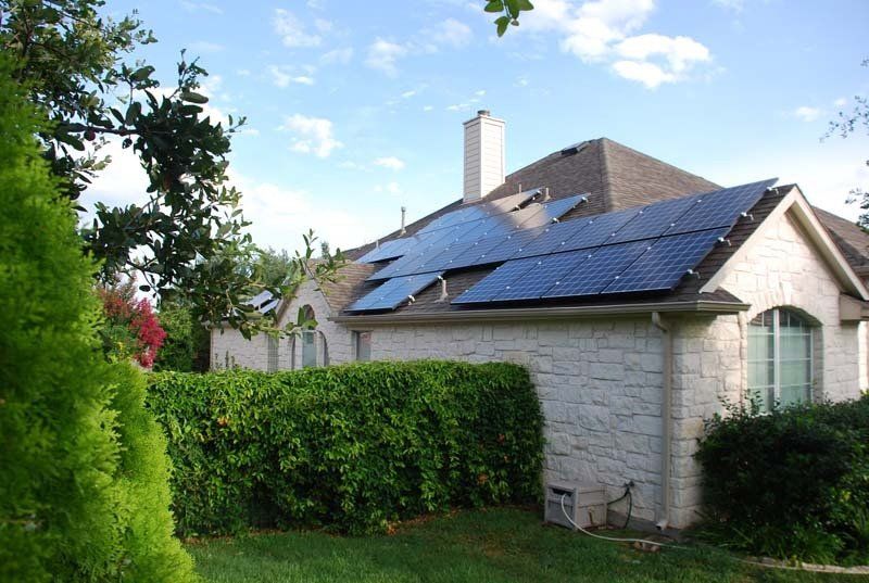 House with solar panels on roof; blue sky, green bushes, white brick exterior.