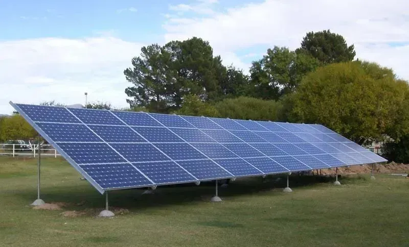 Solar panel array installed in a grassy field, angled toward the sky, with trees in the background.