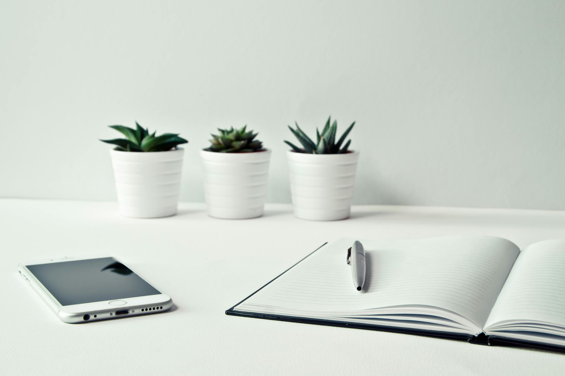 Three potted succulents, a smartphone, and a notebook with a pen on a white surface.