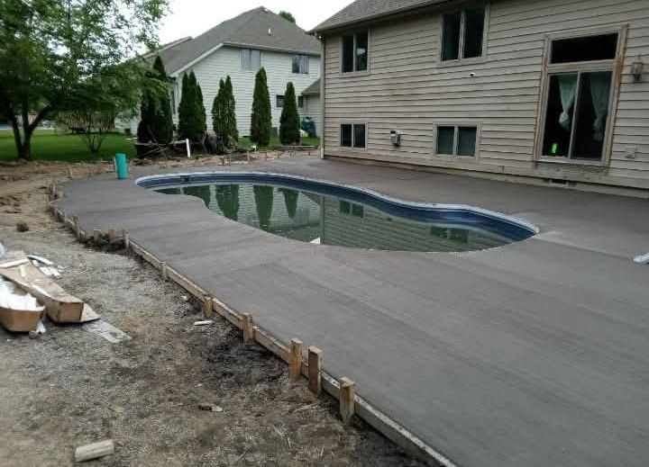 A freshly poured concrete pool deck borders a kidney-shaped pool next to a suburban house under construction.