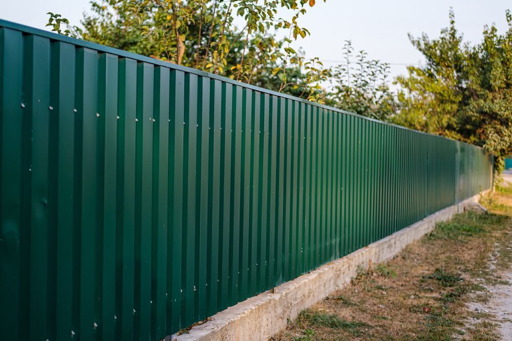 Green corrugated metal fence bordering a grassy area and a dirt path, with trees in the background. — McLuckie Metal Roofing In St George, NSW