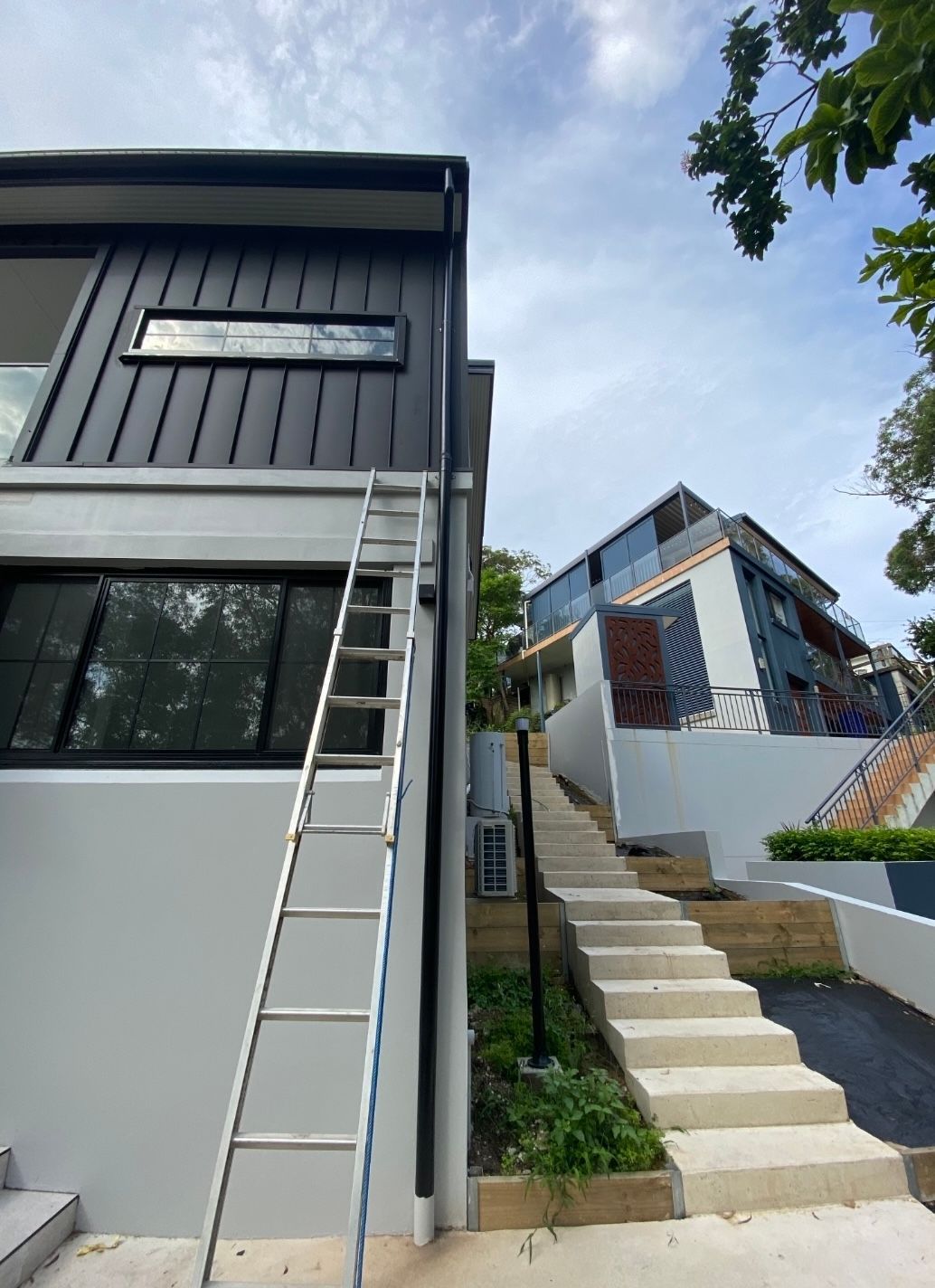 A tall silver ladder leans against a modern two-story house with dark gray siding. Steps lead up a hill to another building. — McLuckie Metal Roofing In Lugarno, NSW
