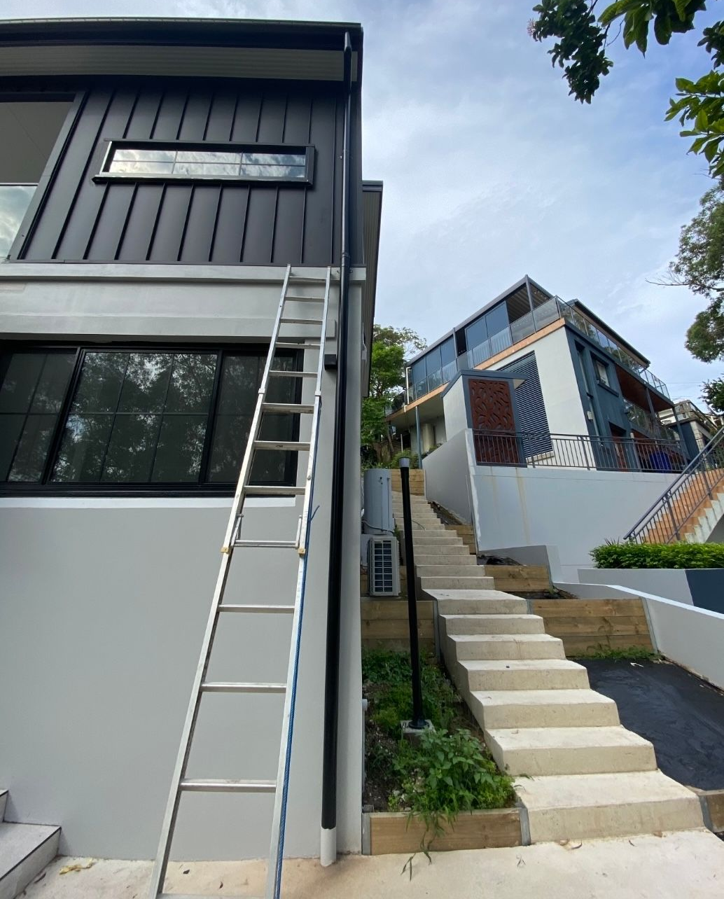 Ladder leaning against a modern two-story building with dark gray siding. — McLuckie Metal Roofing In Lugarno, NSW