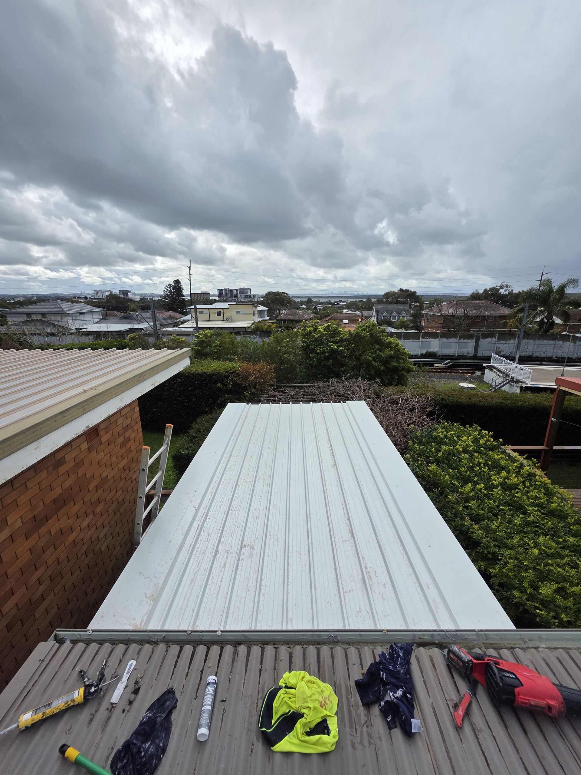 Roofing work in progress; white metal roof sheets against a cloudy sky. Tools and work gloves rest on the roof. — McLuckie Metal Roofing In Lugarno, NSW